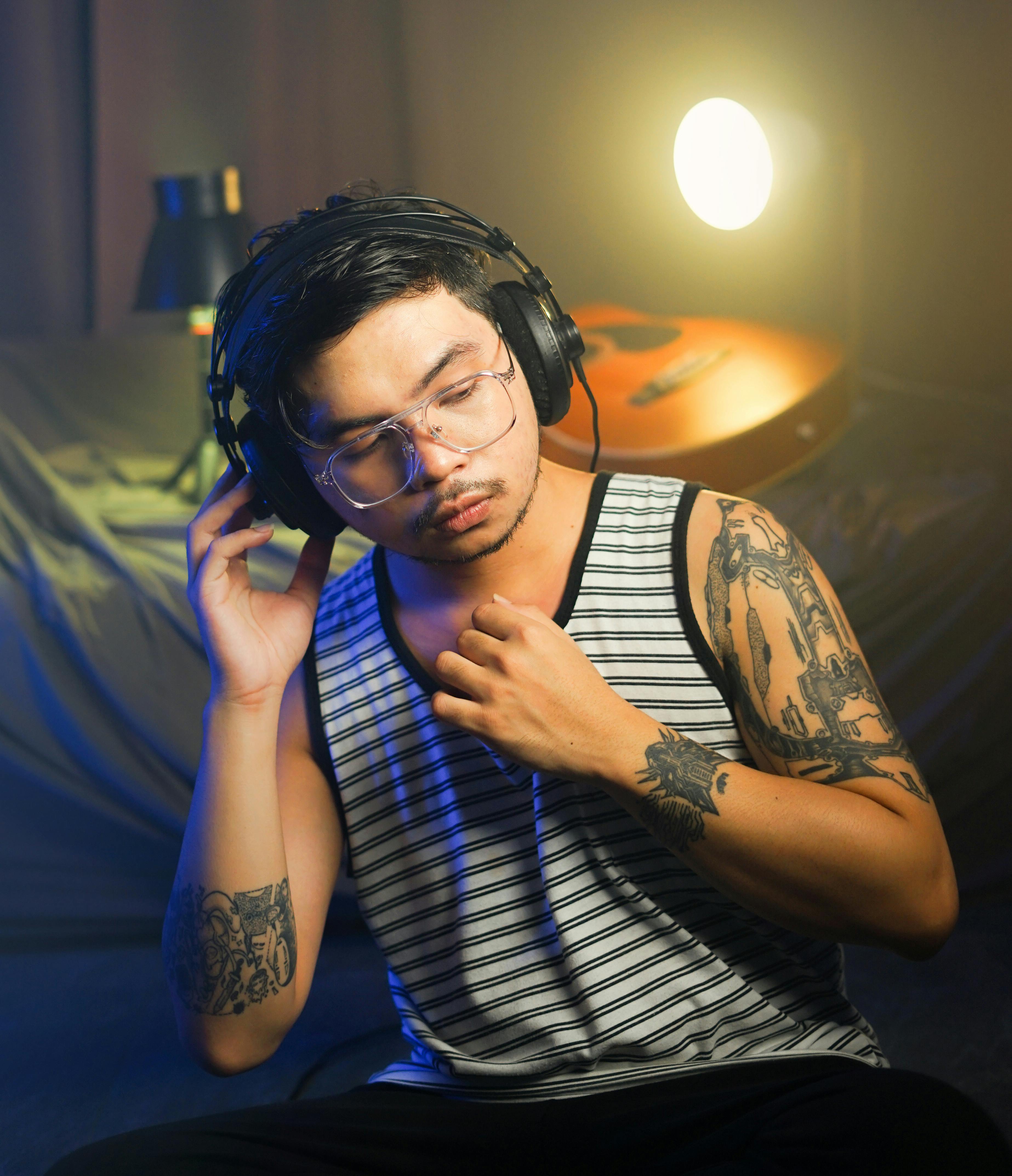 Man with tattoos in a tank top listens to music on headphones in a moody indoor setting.