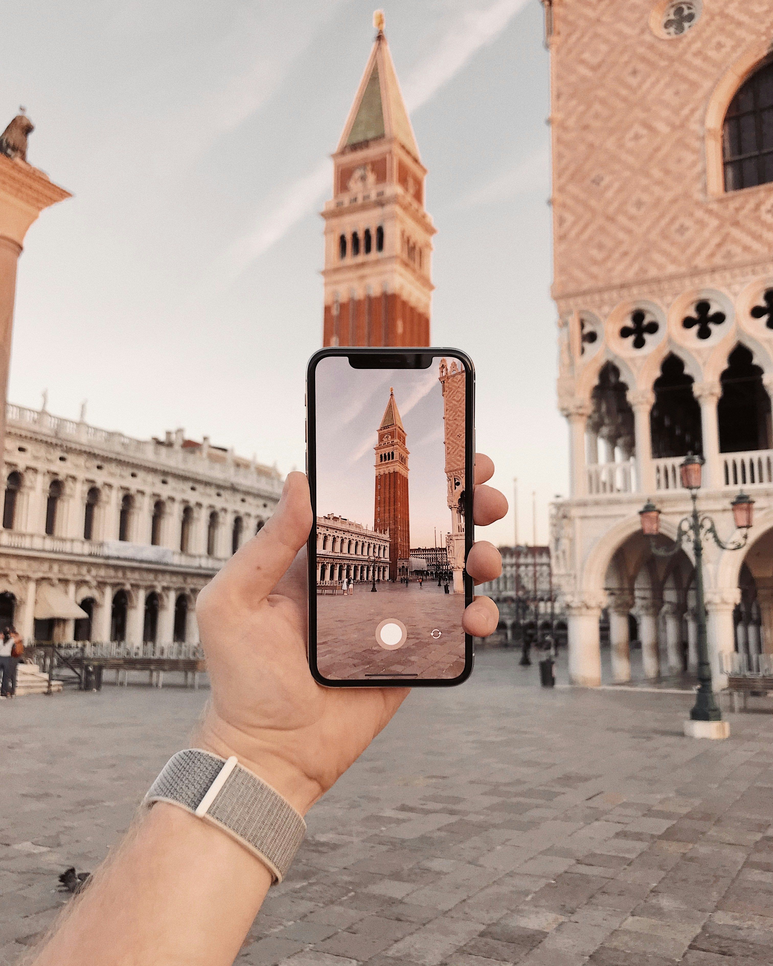 A hand holds a smartphone photographing St Mark's Campanile in Venice, Italy.