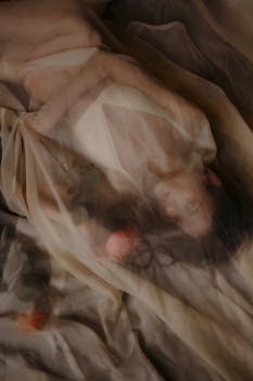 Artistic image of a woman draped in sheer fabric with soft lighting and floral accents.