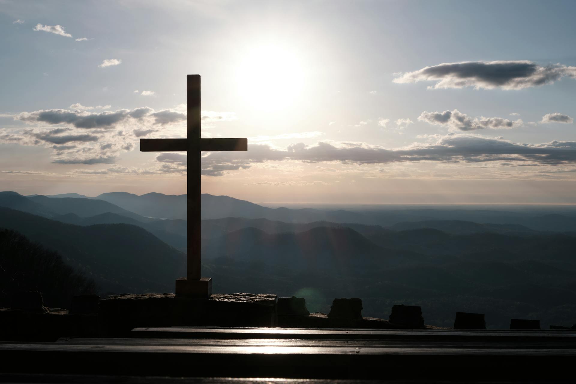Silhouette of a wooden cross against a vivid sunset sky over mountain ranges, with dramatic orange and golden light radiating behind the cross