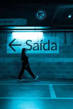 Woman walking in an urban parking lot with a 'Saída' sign, symbolizing exit.
