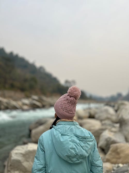 A woman wearing a blue hat and pink jacket stands on rocks near a river