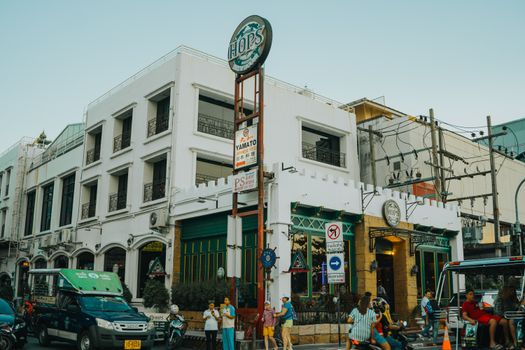 Lively urban street in Bangkok with vehicles and pedestrians near a white building at sunset.