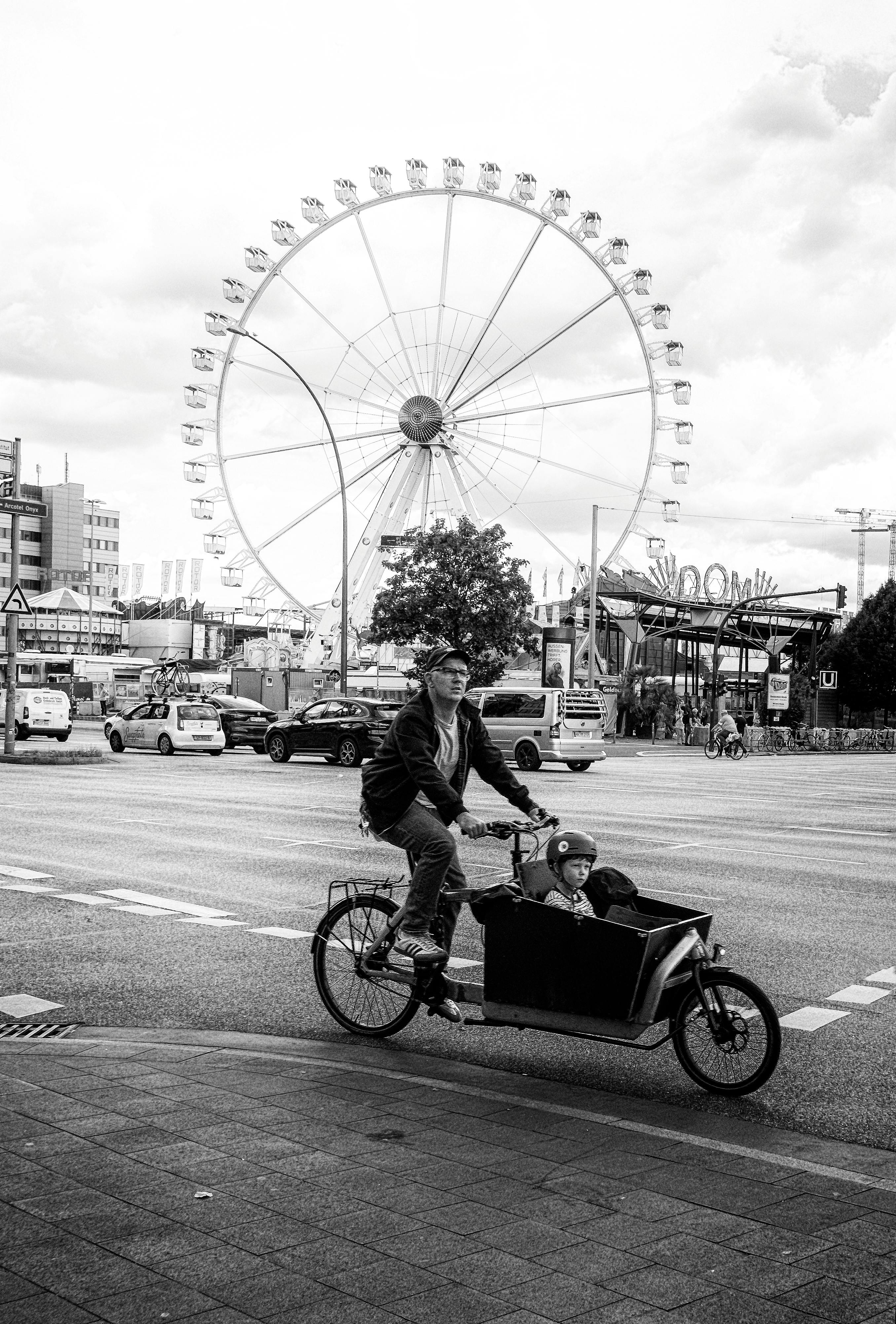 Black and white photo of a father cycling with his son near a city ferris wheel.