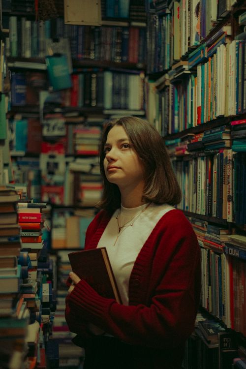 Free A thoughtful woman holding a book stands amidst tall bookshelves in a cozy library setting. Stock Photo