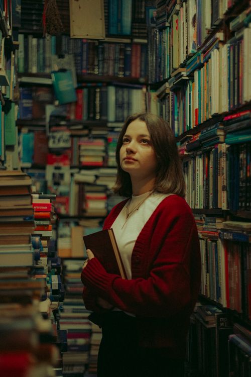 Free A young woman with brown hair stands in a cozy library, holding a book and gazing thoughtfully. Stock Photo