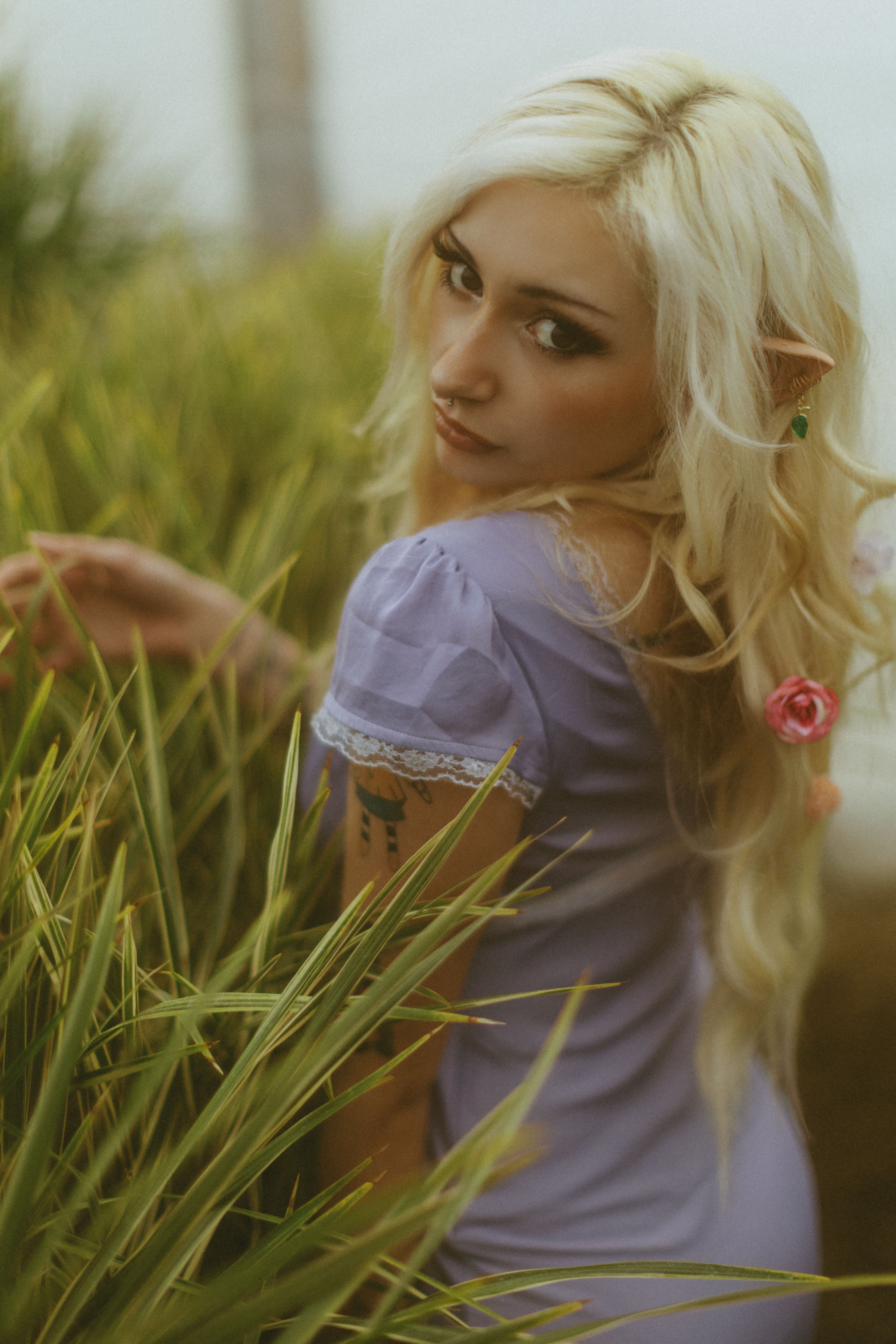 Blonde woman in elf cosplay with lavender dress in a natural setting, evokes a mystical fantasy theme.