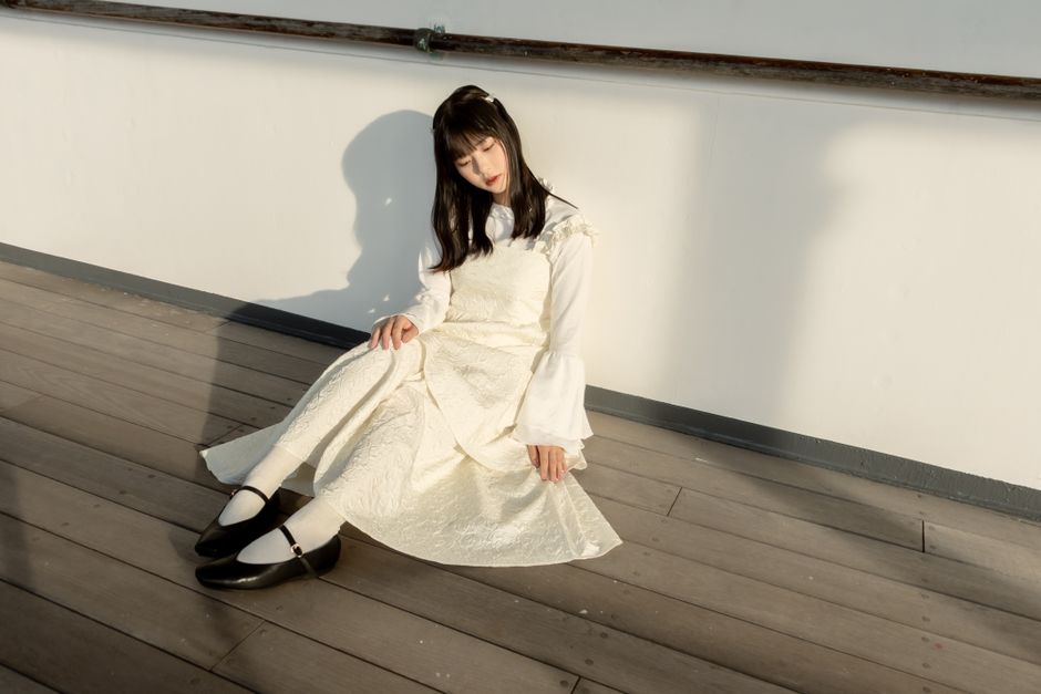 A young woman in an elegant white dress sits peacefully against a wall.
