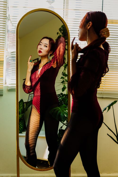 Free Fashion model in red bodysuit posing by a mirror indoors with natural light. Stock Photo