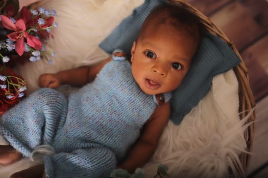 Cute baby in a blue knit outfit lying in a basket with vibrant flowers around.