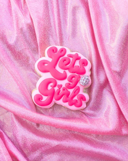 Close-up of a decorative cookie with 'Lets go Girls' text on shimmery pink fabric.