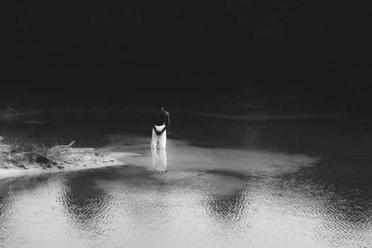 Grayscale photo of person standing alone on water's edge reflecting serenity.