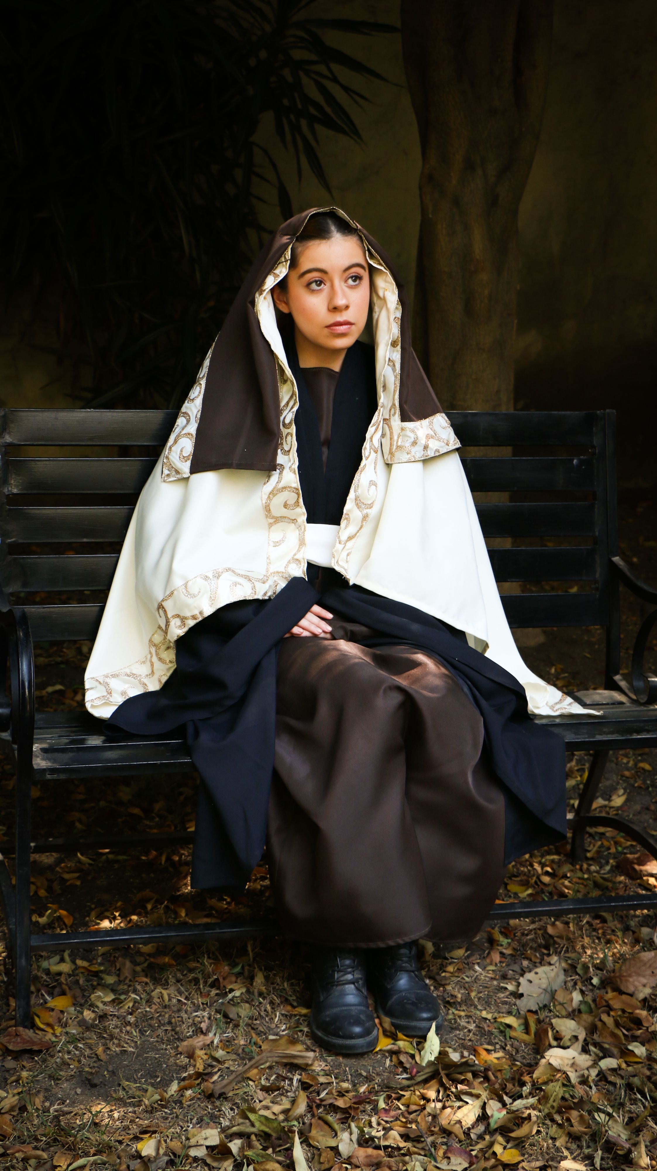 Young woman dressed in a nun costume, sitting on a bench surrounded by autumn leaves.