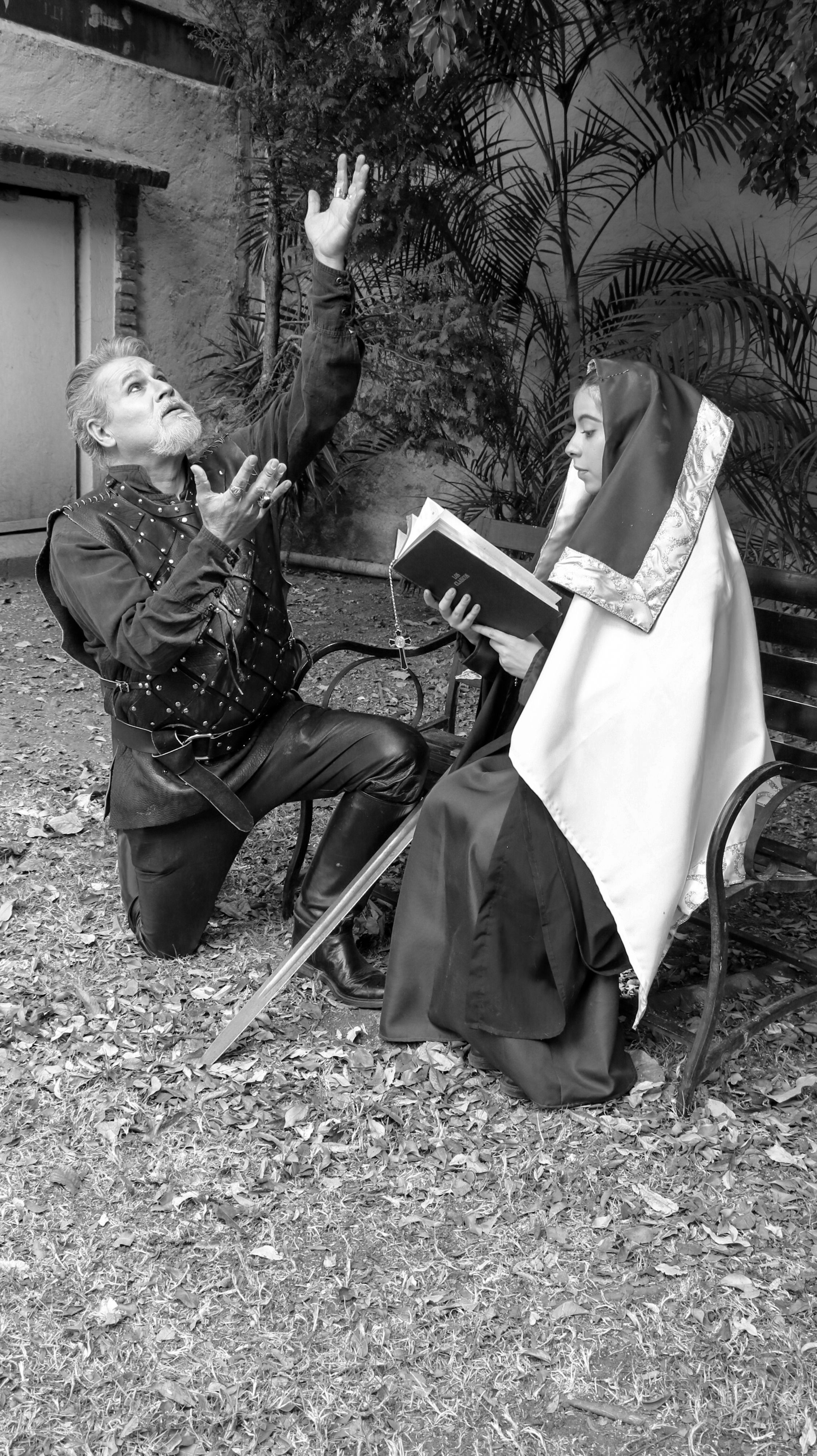 Black and white photo of a kneeling knight and seated nun in medieval attire, outdoors.