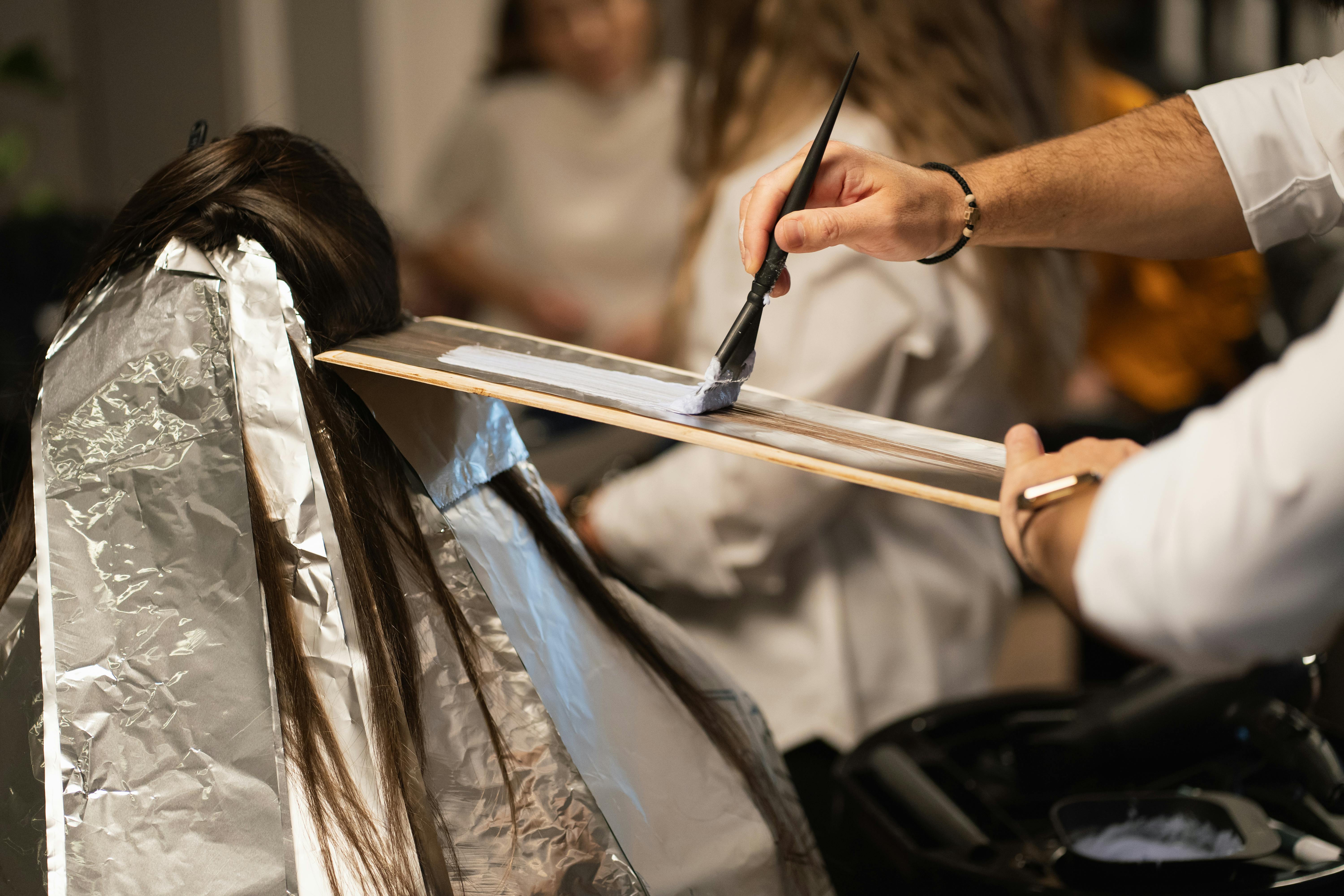 A hairdresser applies color to hair using a brush on a wooden board, with foil wrapped around the hair.