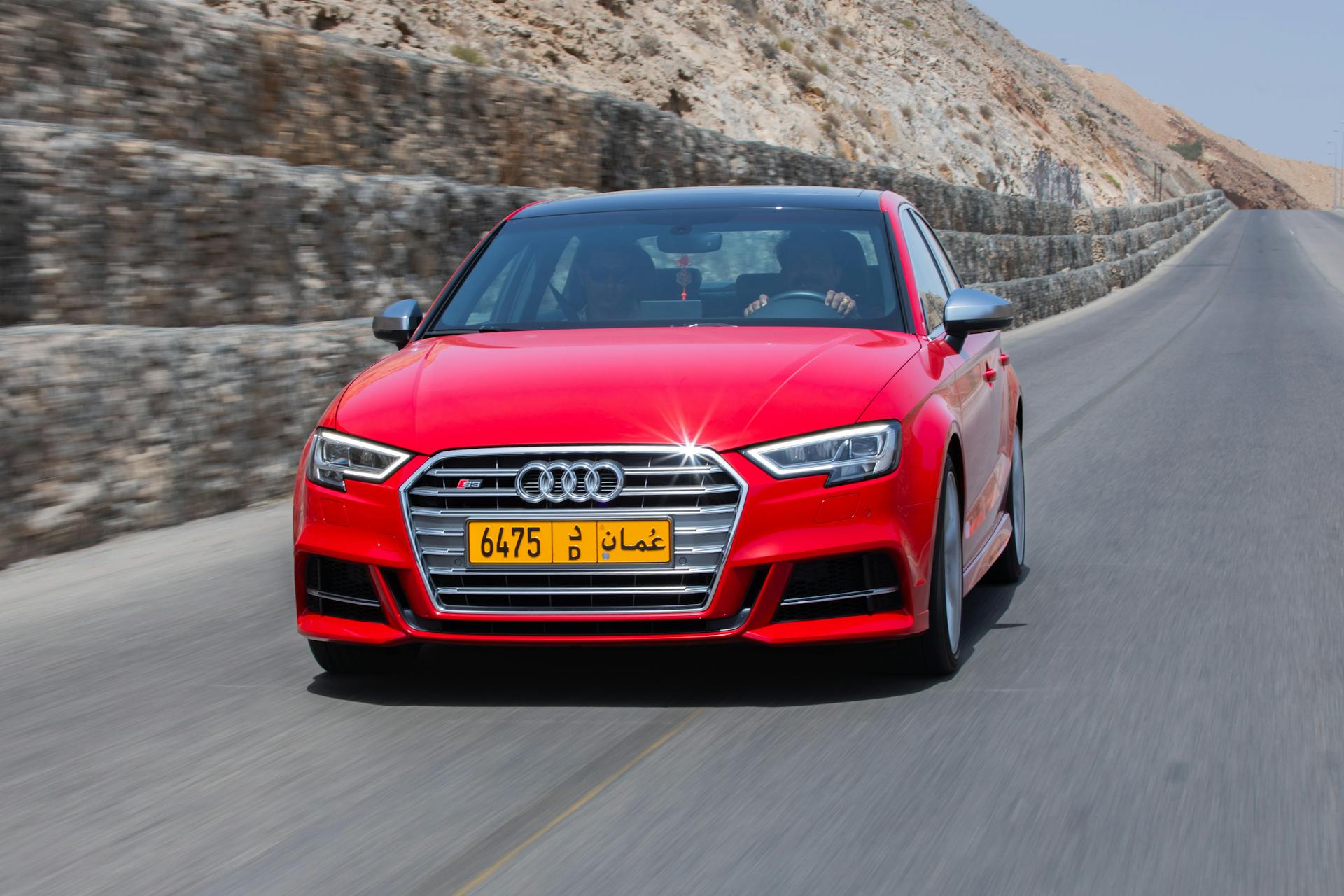 A vibrant red Audi S3 speeds along a scenic mountain road under a clear sky in Oman.