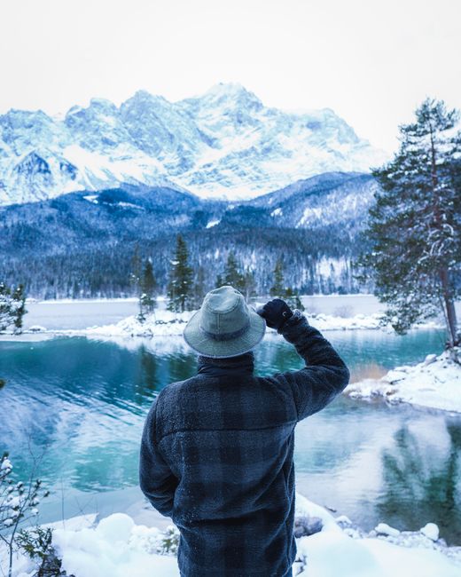 A man in winter attire stands by the scenic Eibsee Lake, gazing at the snow-covered Alps mountains.