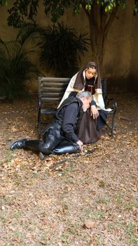 A medieval knight rests on a nun's lap on a park bench surrounded by autumn leaves.