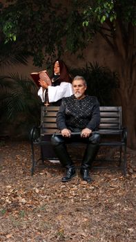 A knight and a nun in medieval costumes sitting on a bench with foliage behind.