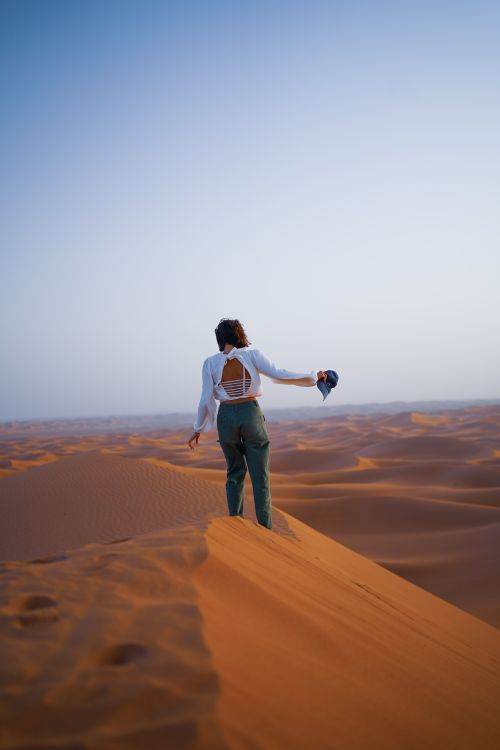 Free A woman stands alone on a desert dune, capturing the vastness of the arid landscape under a clear sky. Stock Photo