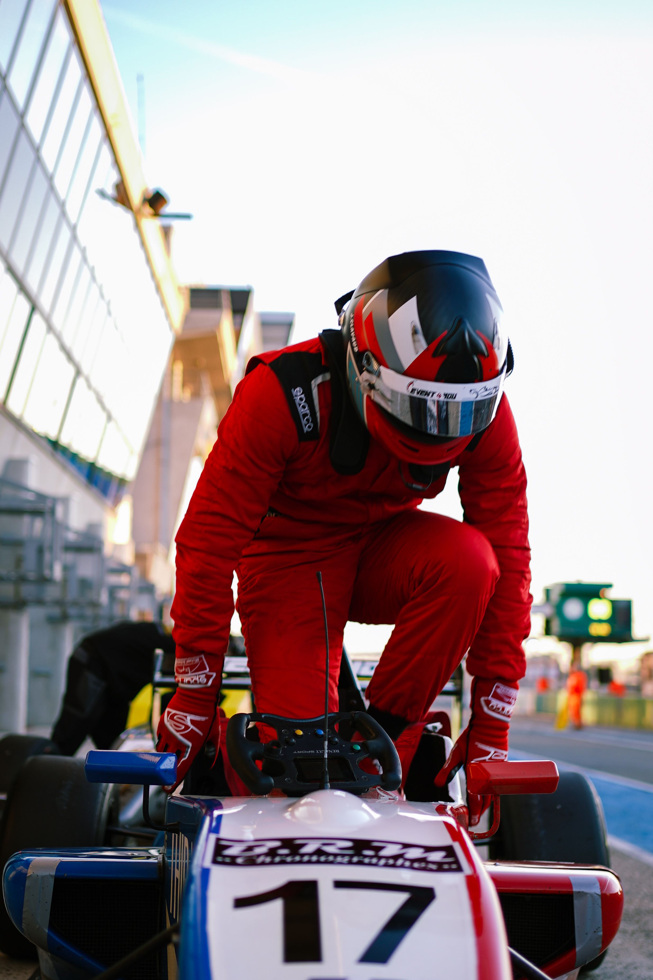 A race car driver in red gear prepares to enter a Formula 3 car at a racing circuit with modern architecture.
