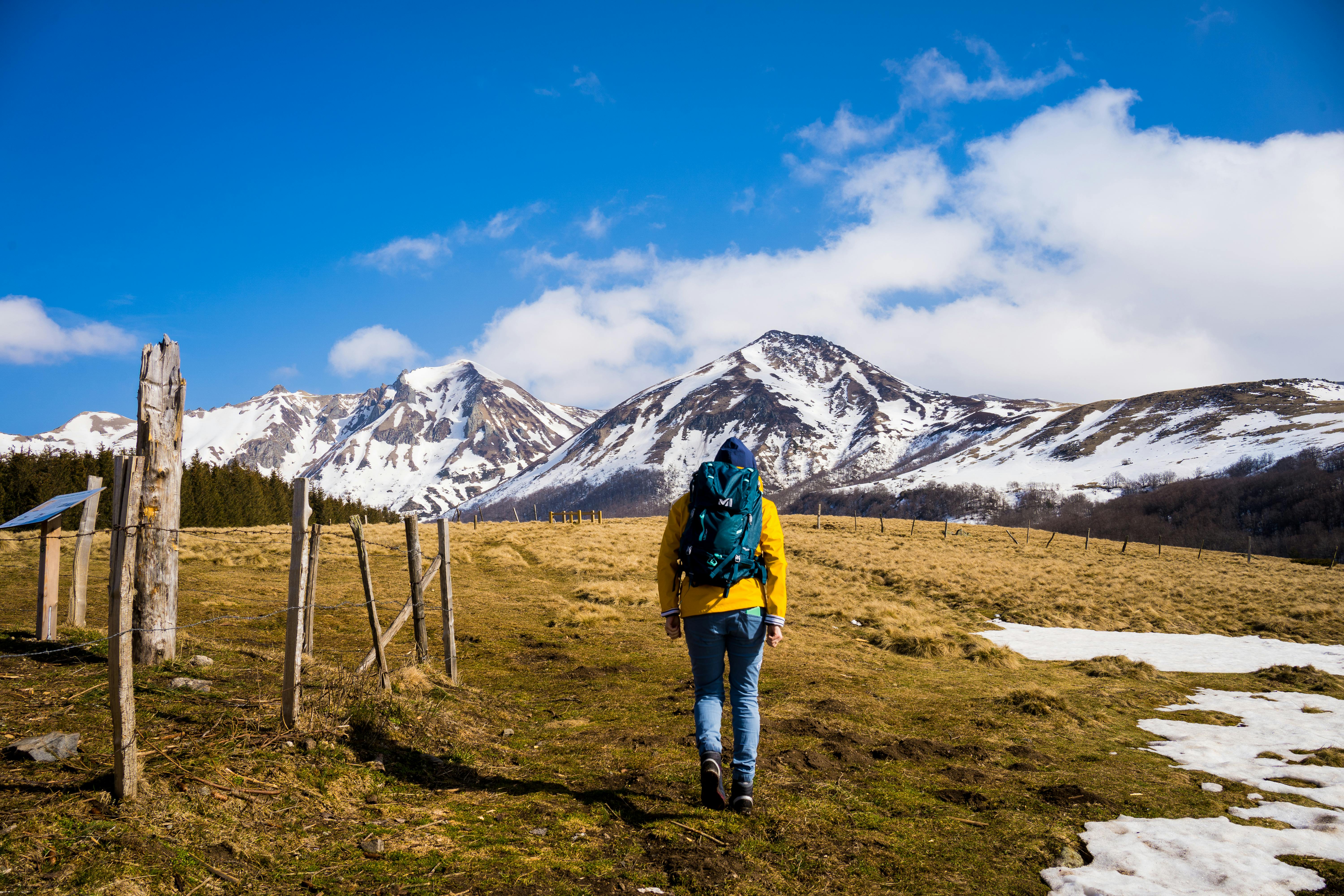 A hiker with a backpack walks towards snow-covered mountains on a sunny day.