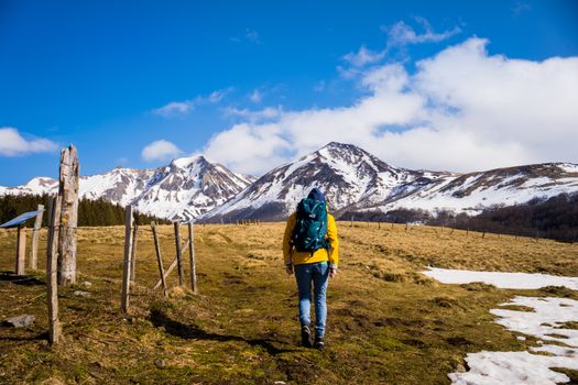 A hiker with a backpack walks towards snow-covered mountains on a sunny day.