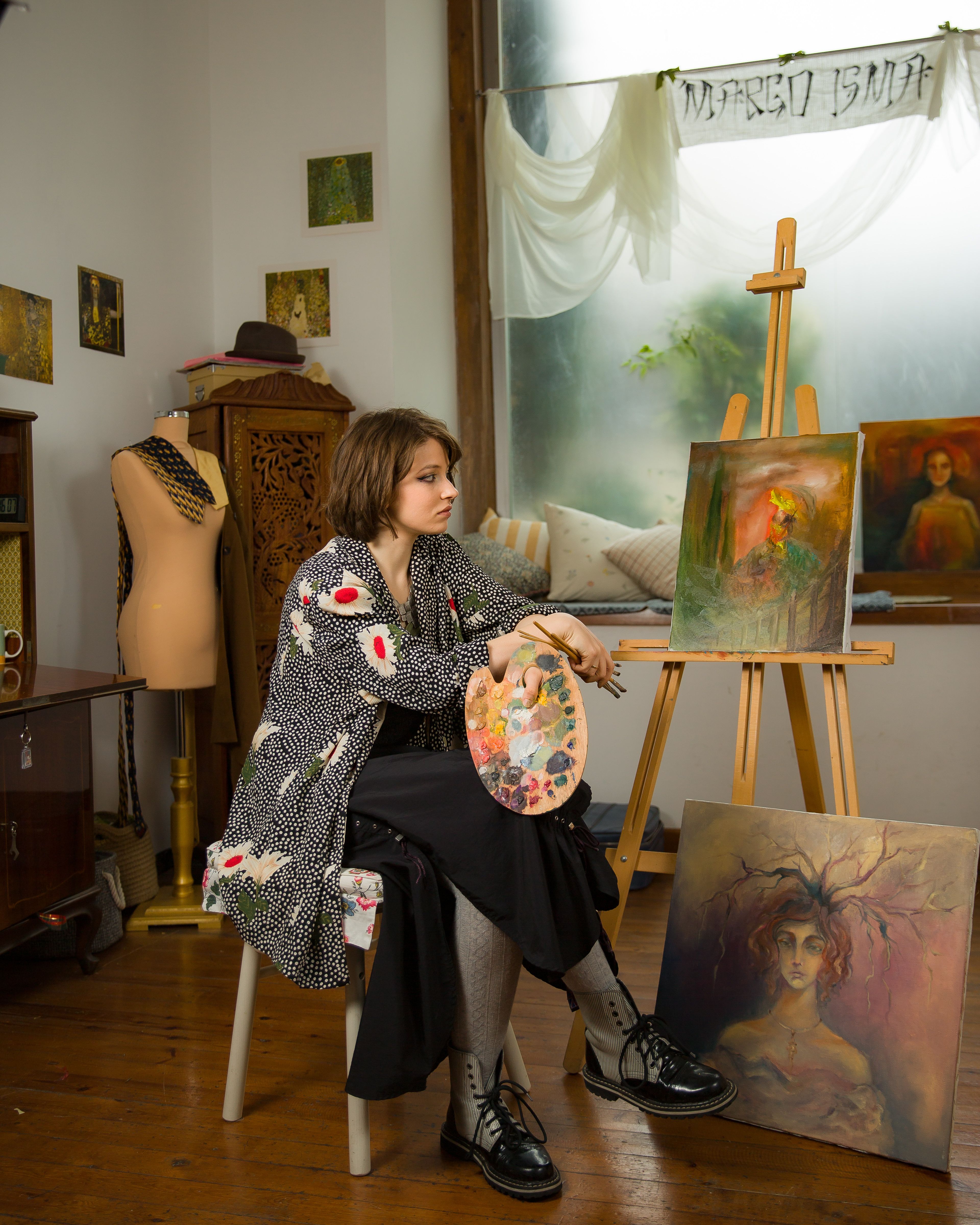 Woman artist sitting in a studio with her paintings and palette, showcasing creativity.