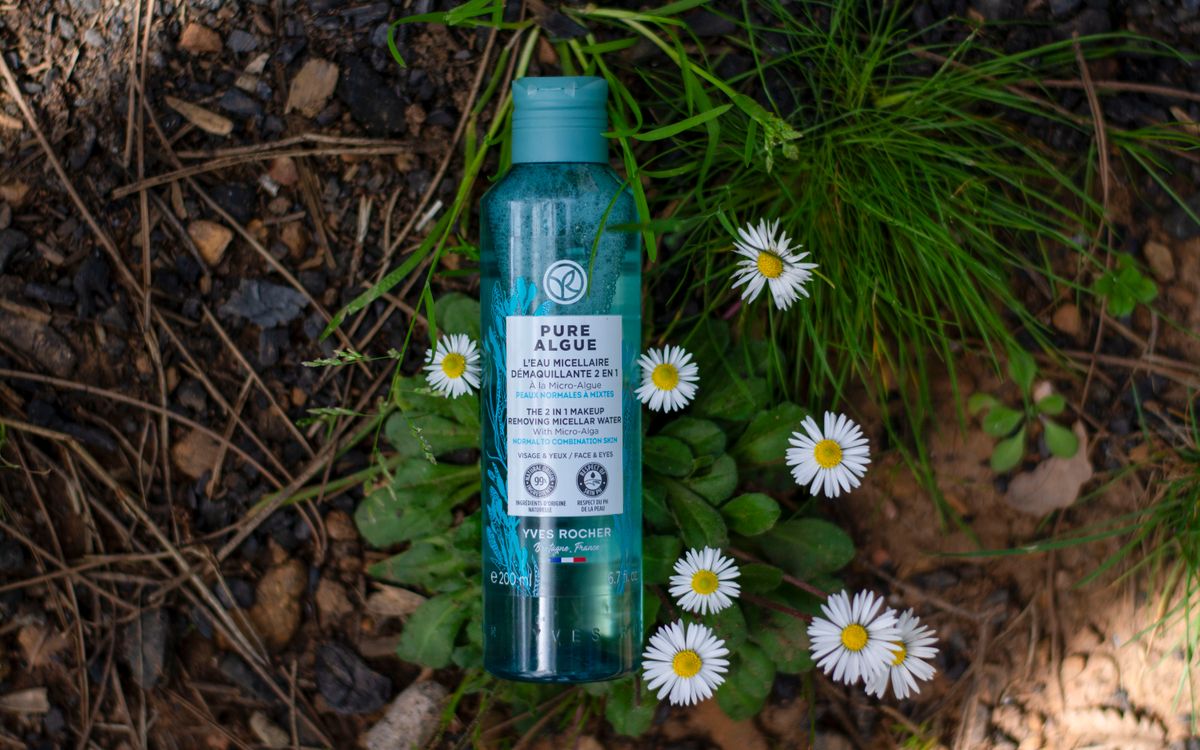 A bottle of micellar water surrounded by daisies on a natural ground surface.