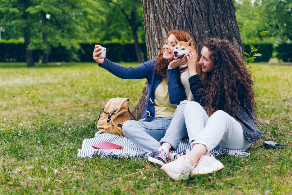 Two women and a Shiba Inu dog enjoying a sunny day in the park while taking a selfie together.