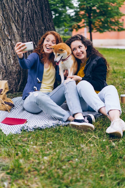 Two women sitting on the grass taking a selfie with their dog