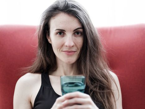 Portrait of a young woman sitting indoors, enjoying a refreshing glass of water on a casual day.