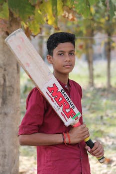 Teenager holding a cricket bat in a sunny park, showcasing sporty confidence.