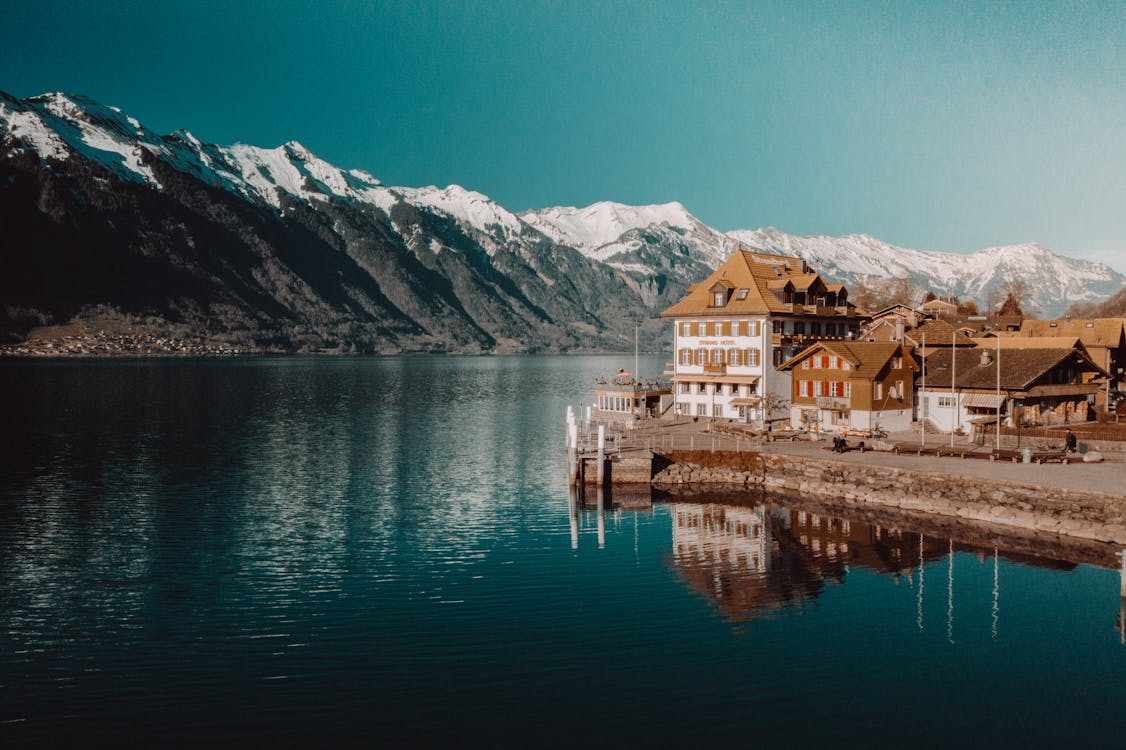 Edificio De Hormigón Blanco Y Marrón Junto Al Cuerpo De Agua Cerca De Las Montañas Rocosas Cubiertas De Nieve Bajo Un Cielo Azul Claro