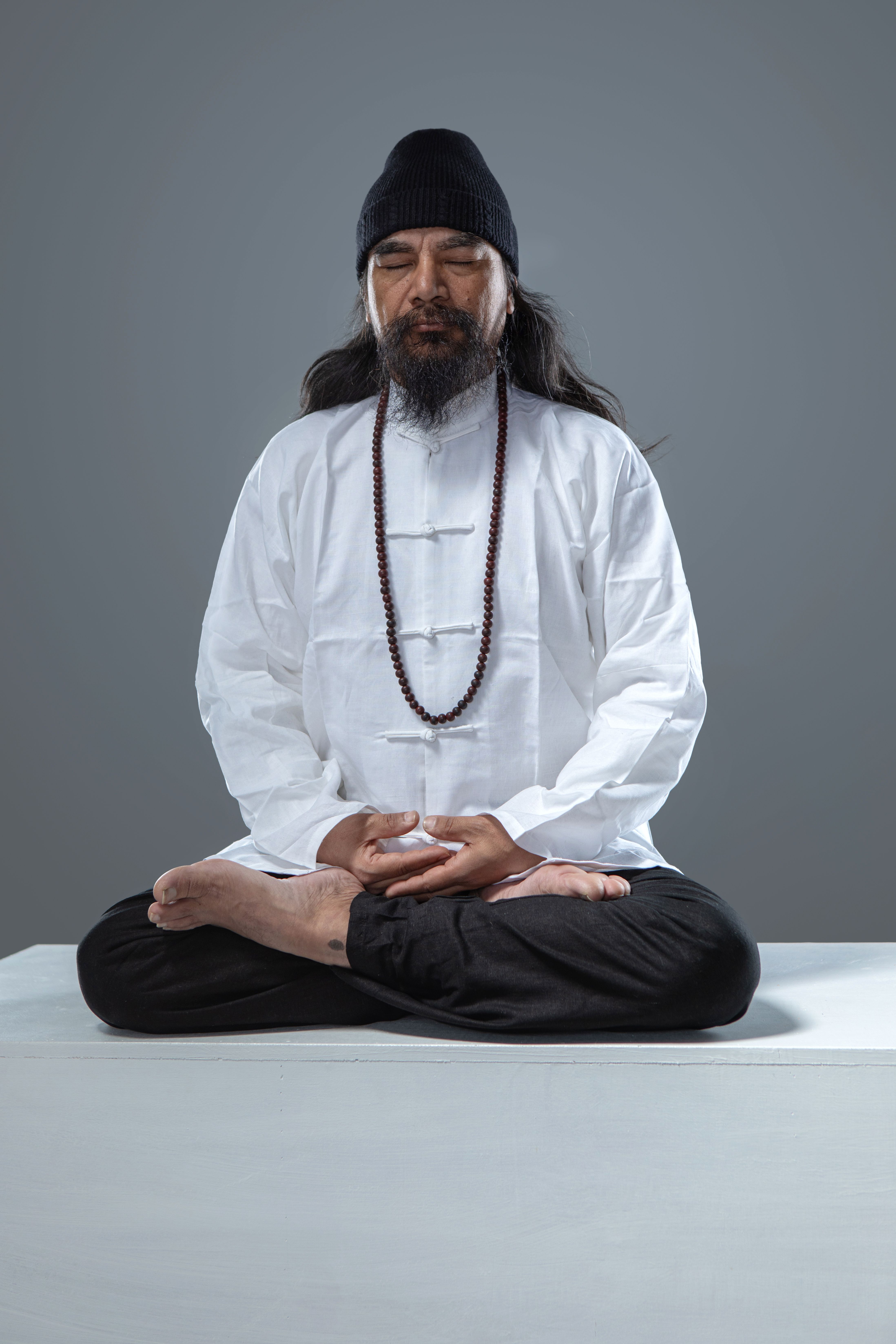 Man wearing traditional attire meditates cross-legged in a studio.