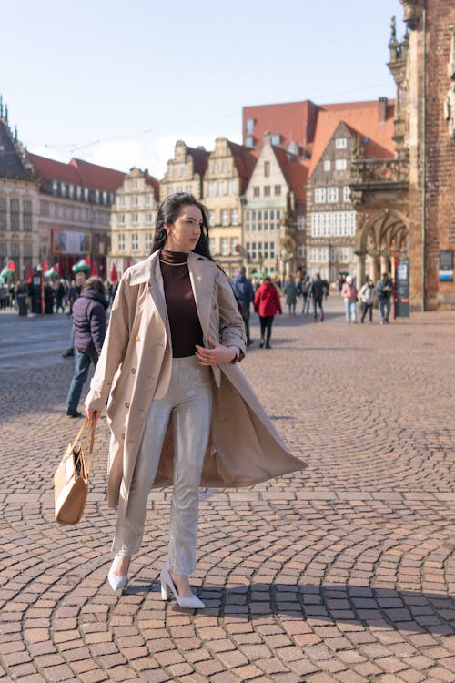 A woman in a trench coat and white pants walking down a cobblestone street