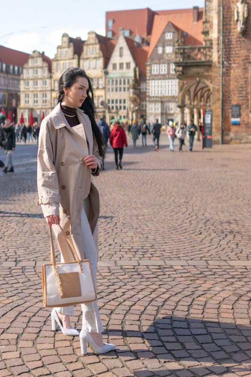 A woman in a white coat and beige bag walks down a cobblestone street