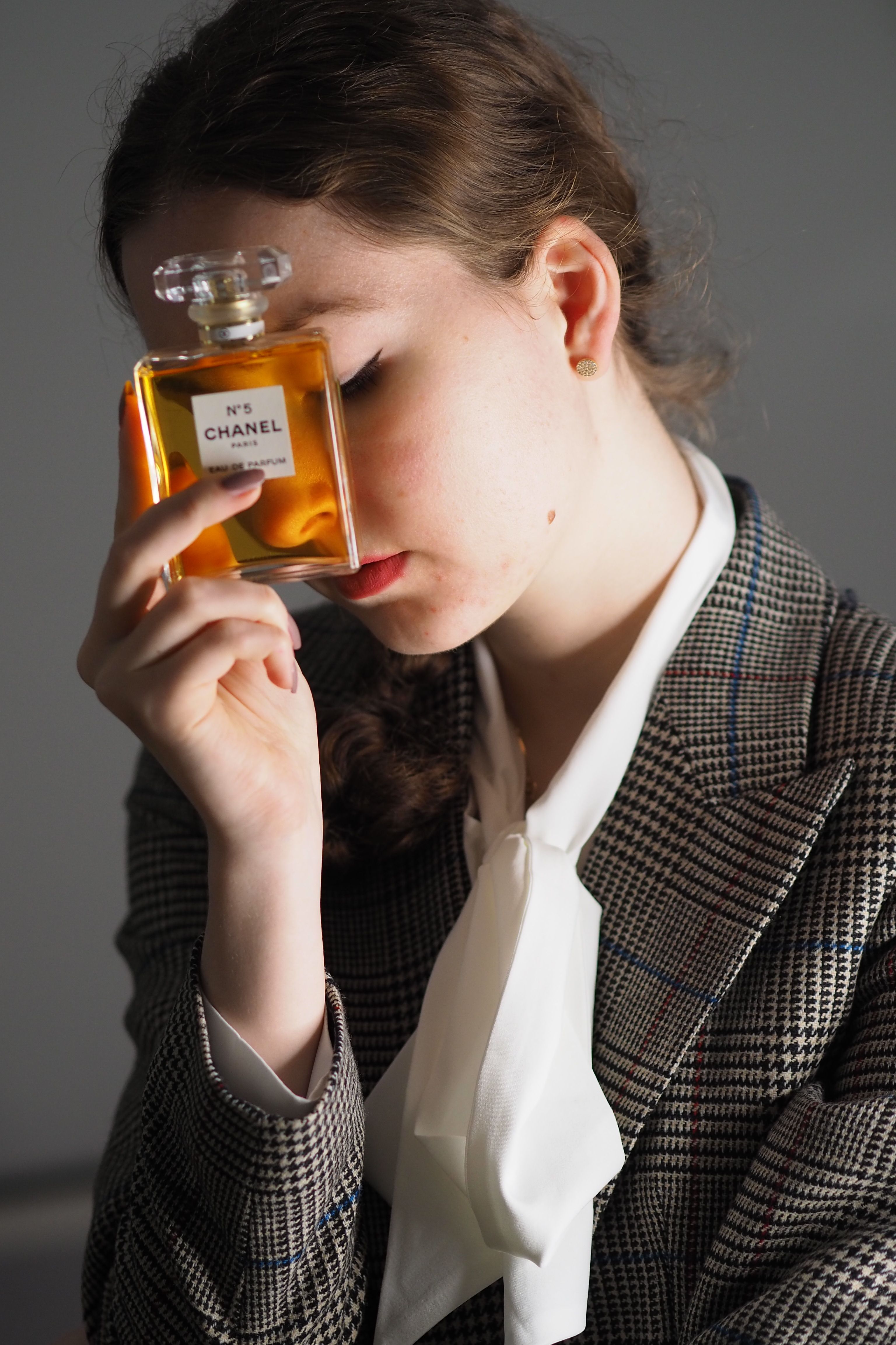A young woman in a checked jacket holds a Chanel perfume bottle against her face, eyes closed.