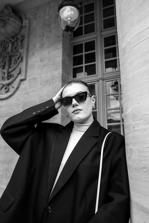 A woman in a black coat and sunglasses leaning against a wall