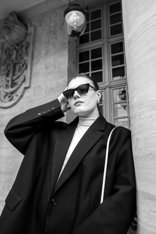 A woman in sunglasses and a black jacket leaning against a pillar