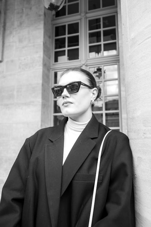 A woman in sunglasses and a black coat leaning against a wall