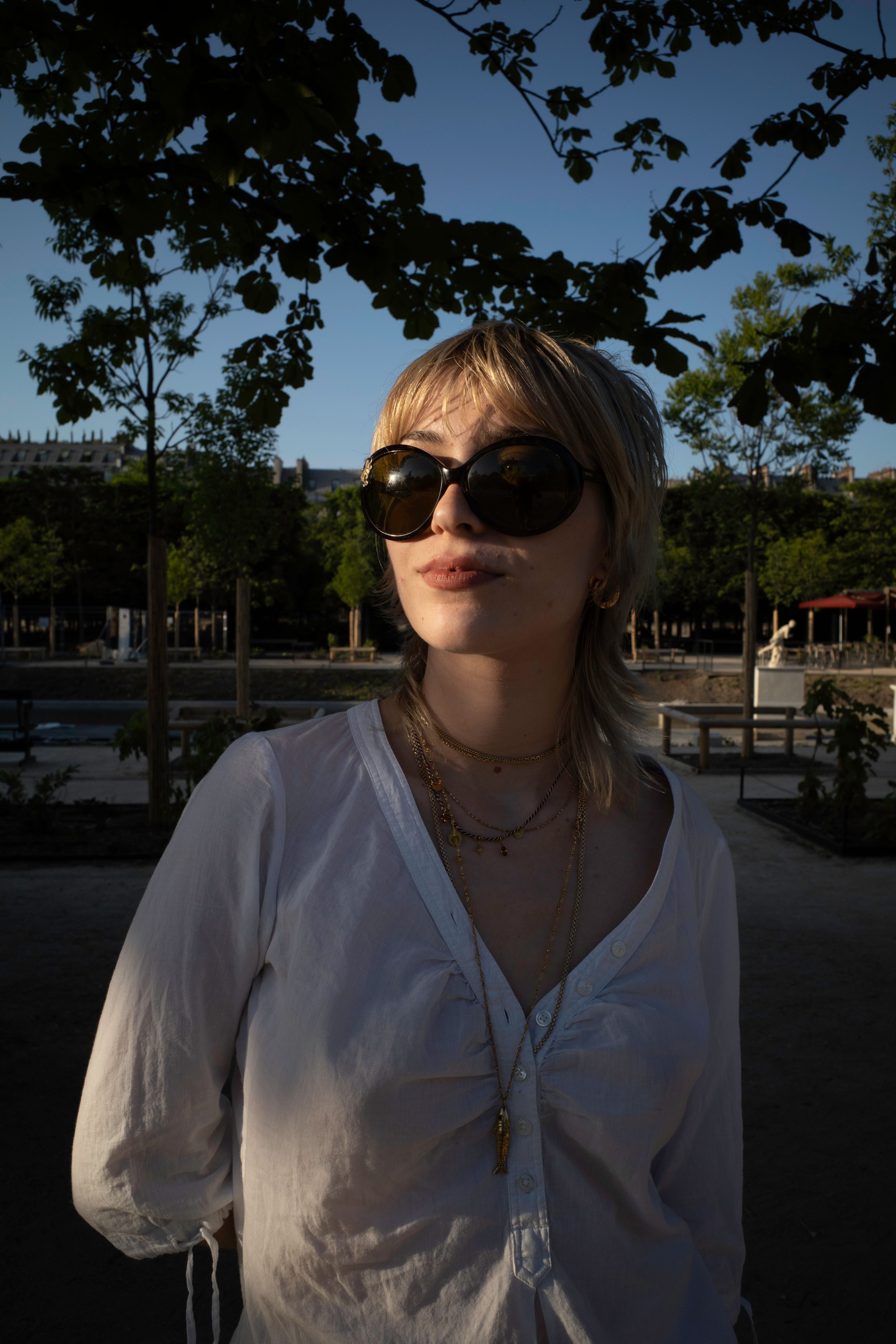Caucasian woman with sunglasses and necklaces posing outdoors in a sunny setting with trees.