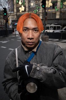 Asian man with orange hair standing confidently on city street, showcasing modern urban fashion.