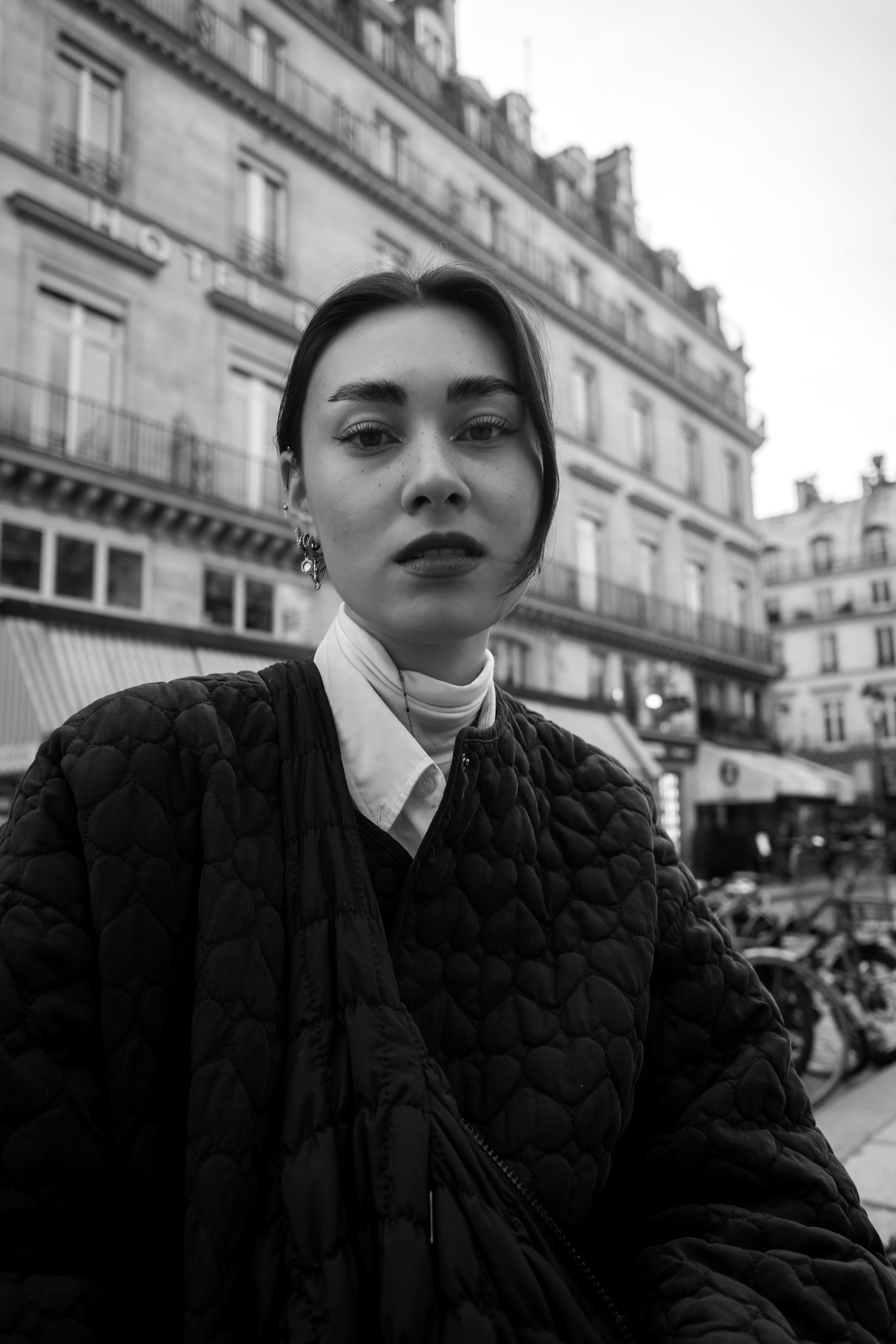 Stylish woman in a quilted jacket poses outdoors in a city setting, captured in black and white.