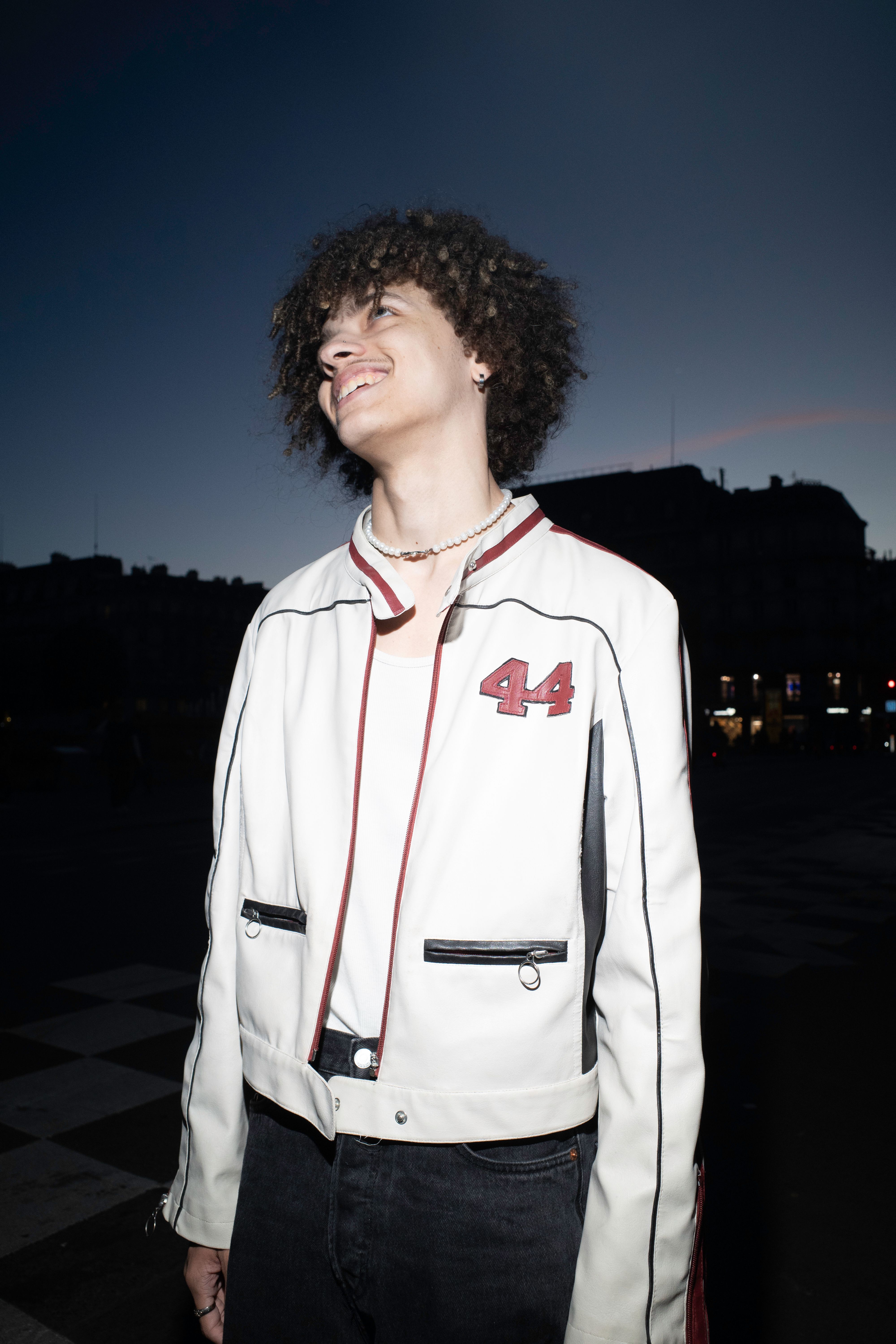 Smiling young man in a fashionable white jacket with '44' detail at dusk in an urban setting.