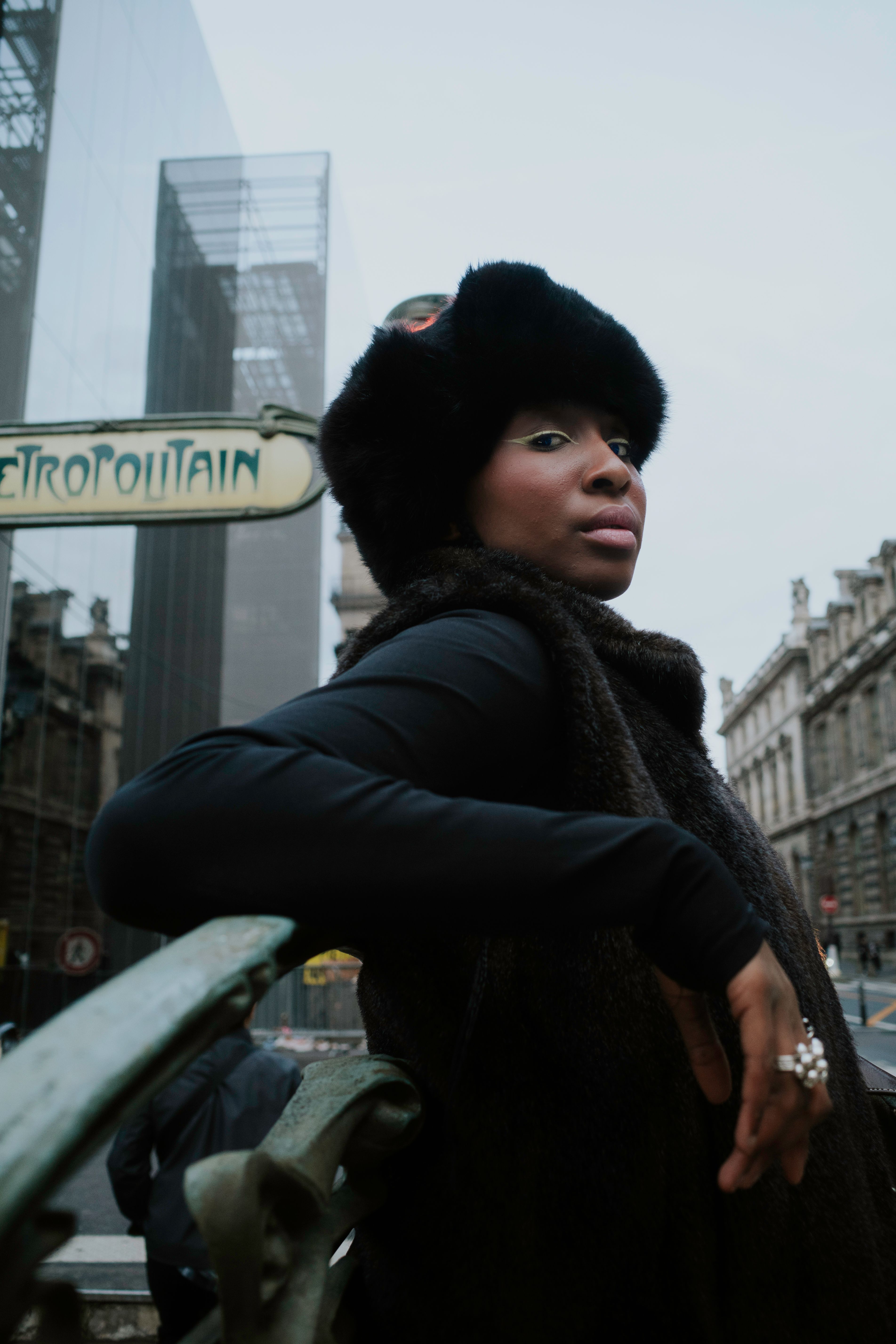 A fashionable woman in a fur coat poses confidently near urban architecture.