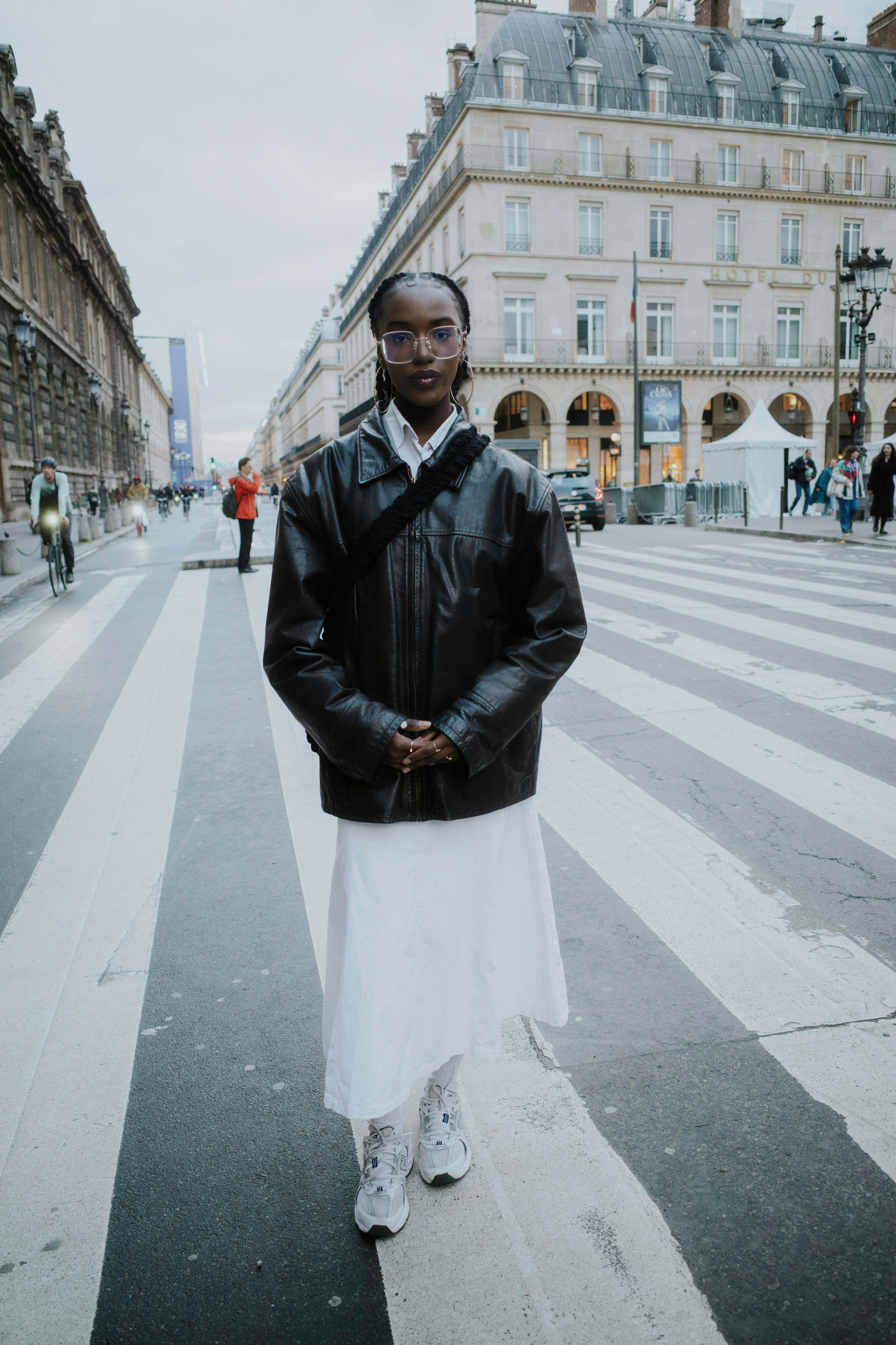 Stylish woman poses in a city crosswalk wearing a leather jacket and white skirt.