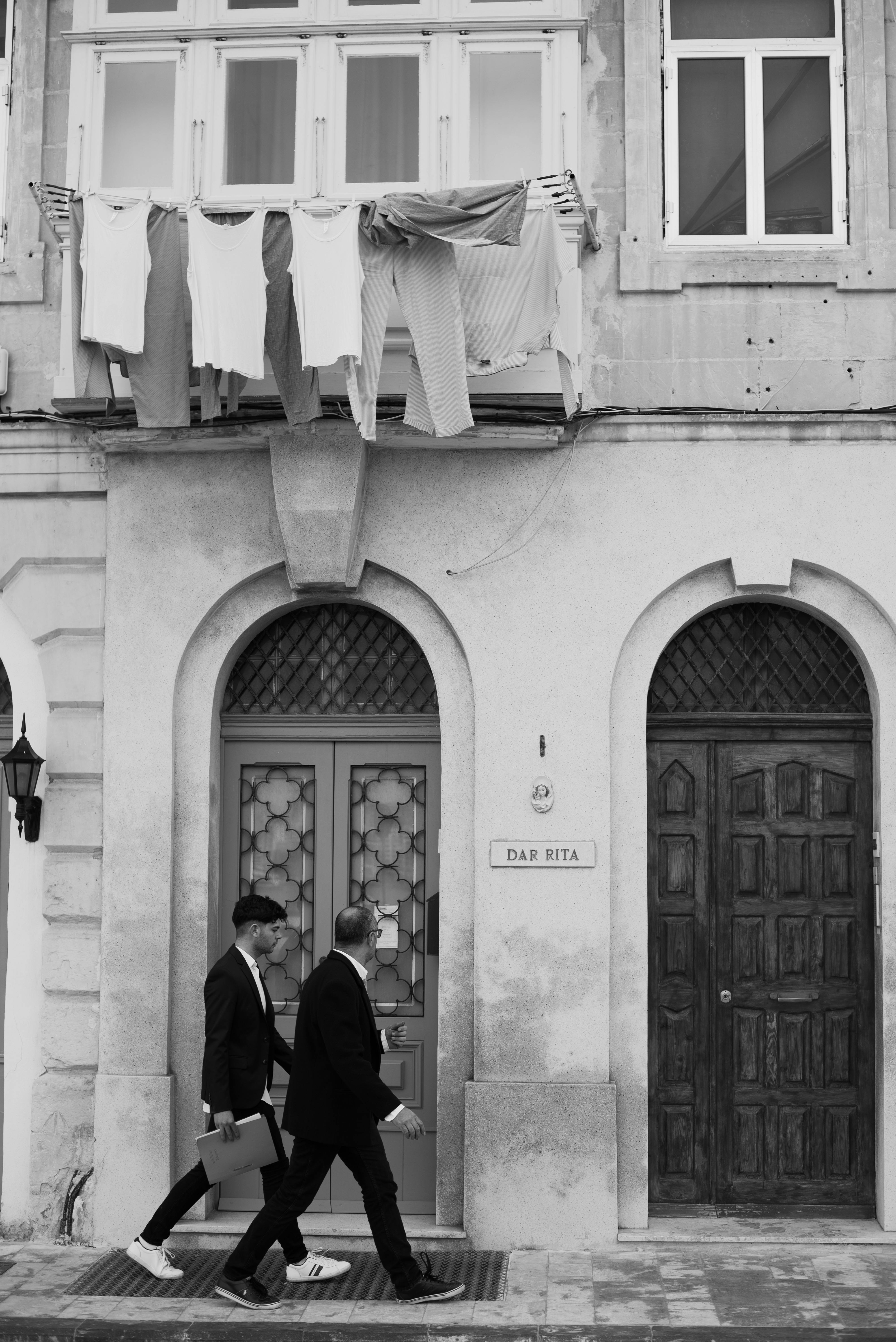 Black and white photo of two men walking past a building with laundry drying.