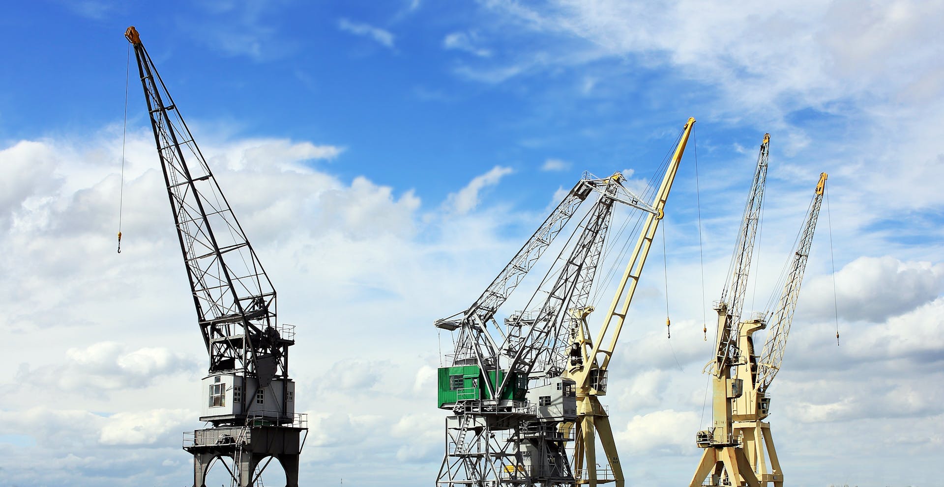 Industrial cranes reaching into a bright blue sky with scattered clouds, a symbol of construction and industry.