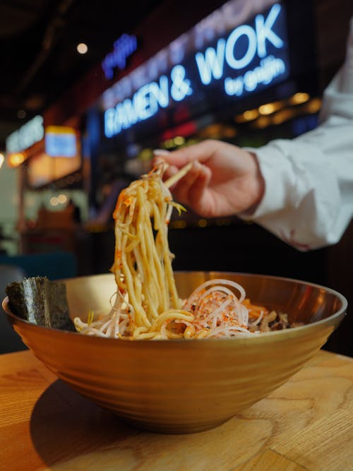 A person holding a bowl of noodles in front of a wooden table